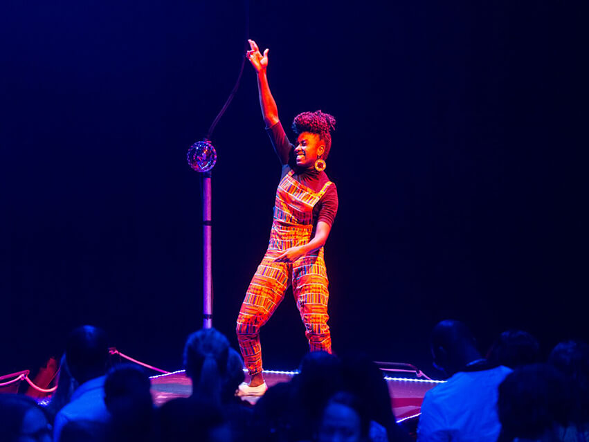 An African British female wearing orange dungarees with colourful rectangular patterns raises her arm powerfully on the dance floor, the crowd watch on and surround her.