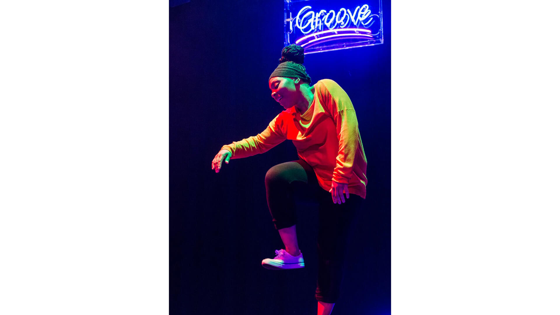 A black woman wearing an orange top and grey leggings moves rhythmically on the dancefloor. Above her is a neon sign that hangs from the ceiling that reads: “Groove”.