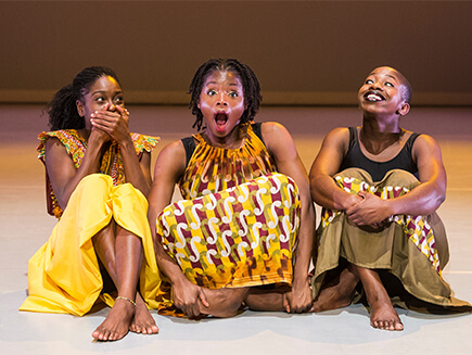 Three black women sit playfully on set, Sistofe with her hands covering her mouth, Habibat with her mouth wide open and a smiling Shanelle with arms crossed.