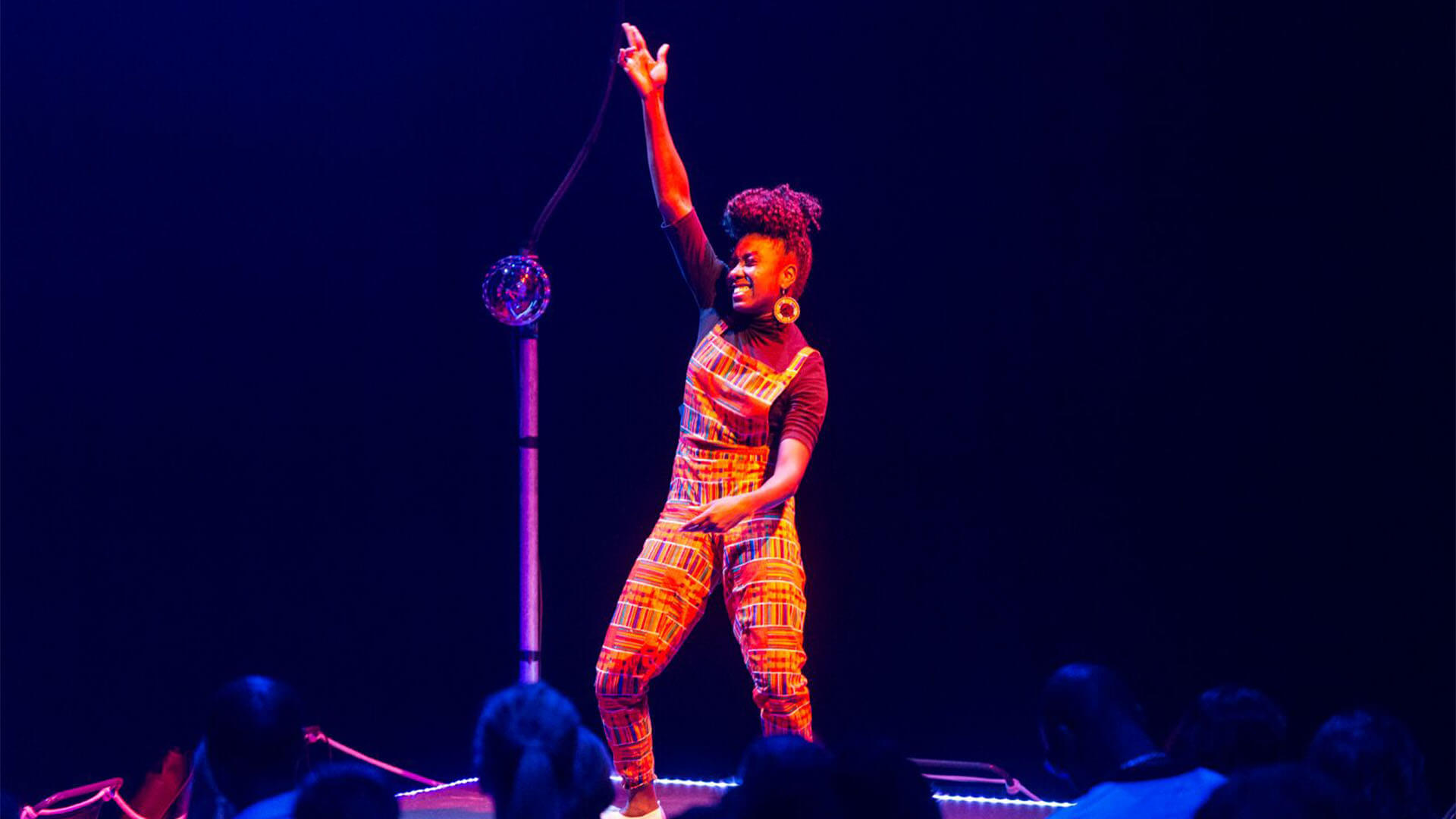 An African British female wearing orange dungarees with colourful rectangular patterns raises her arm powerfully on the dance floor, the crowd surround her.