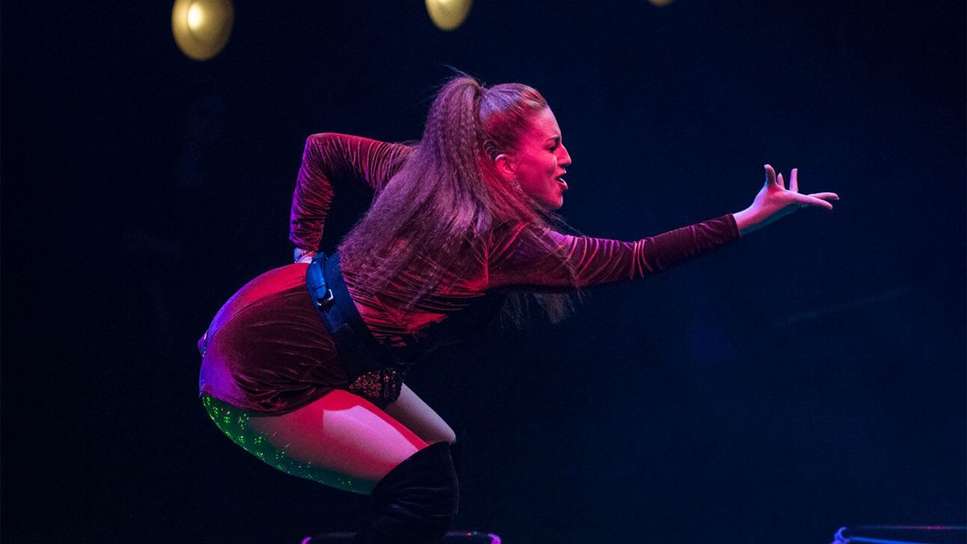 A white woman wearing a red velvet jumpsuit, knee-high boots, long ginger hair held in a high ponytail with one arm extended out in slow motion.