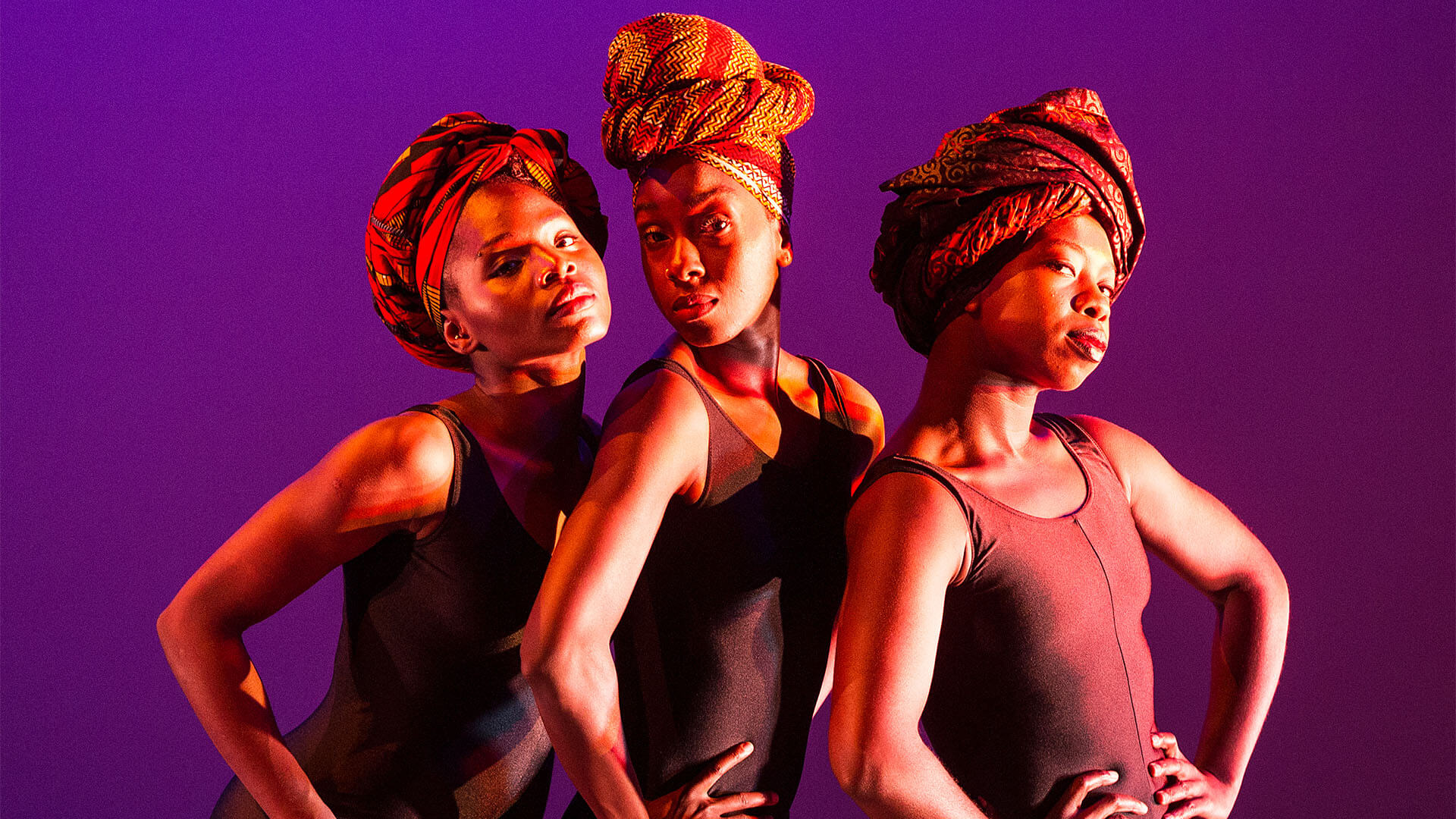 Three black women pose and look confidently at us with their hands on their hips. The stage is lit with warm, purple light.