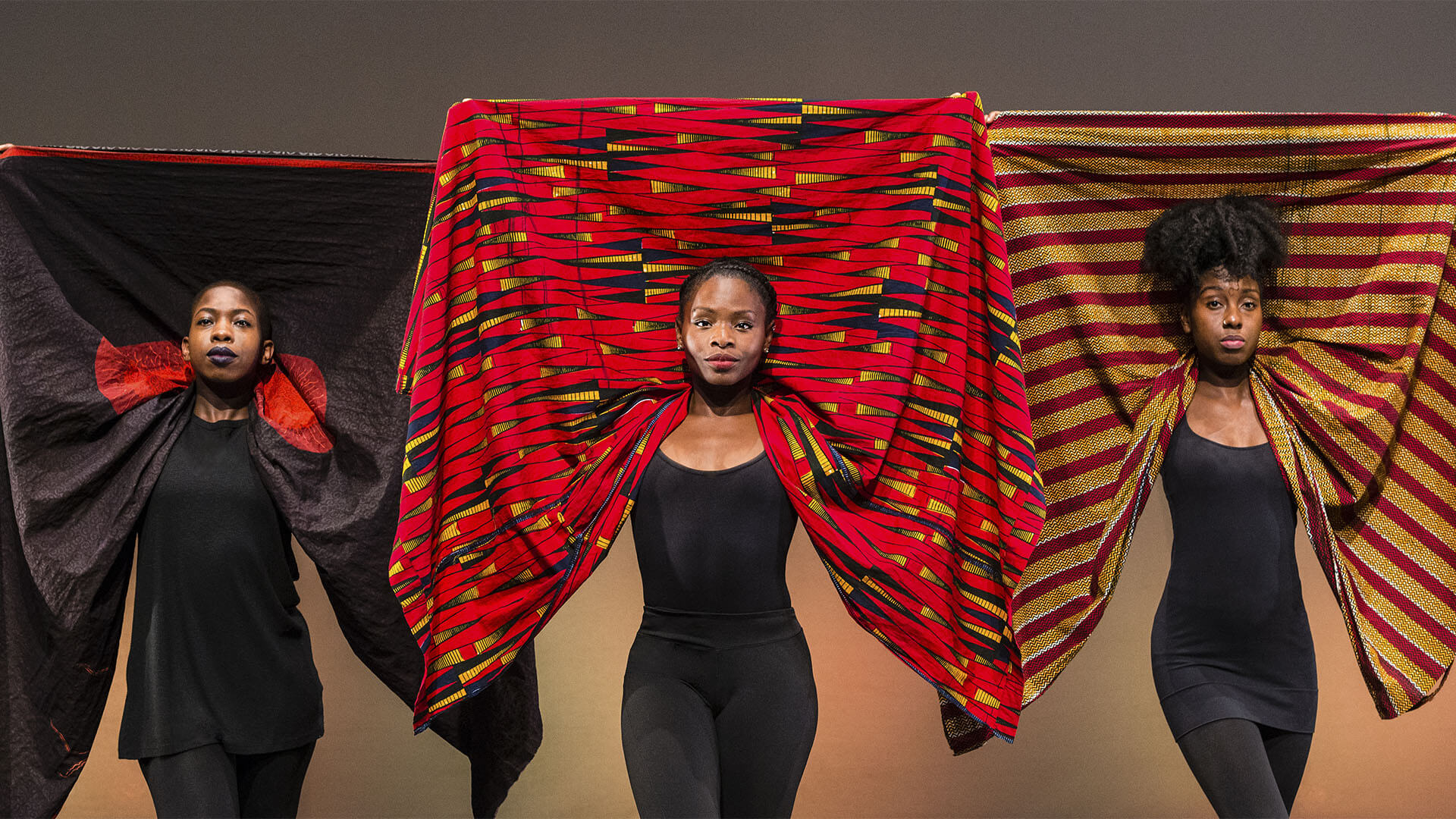 Three black women hold their head wraps up behind their heads. The three head wraps are black with hints of red, predominantly red with black and yellow triangles and striped red and mustard.