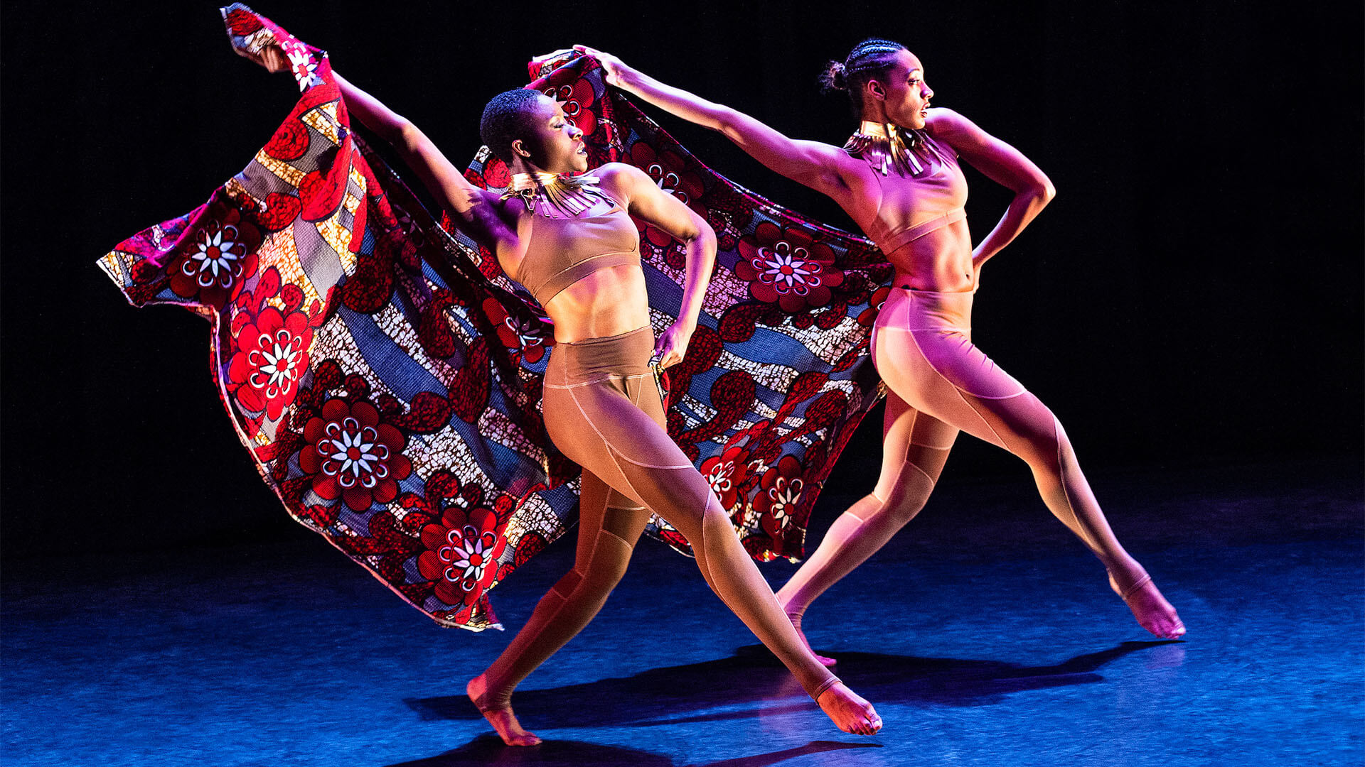 Two black women stride confidently on stage with their colourful burgundy, blue and red head wraps held powerfully behind them.