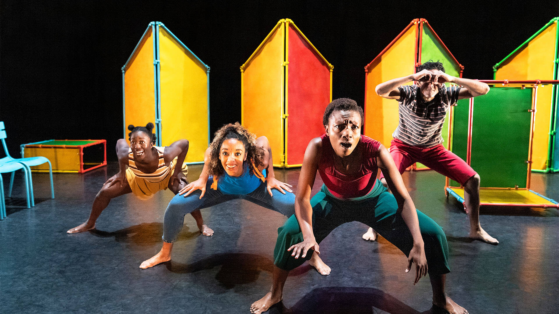 Four performers group together on set which combines different pastel colours: yellow, red, orange, light green, light blue, dark green. It looks a little bit like a kids’ playground.