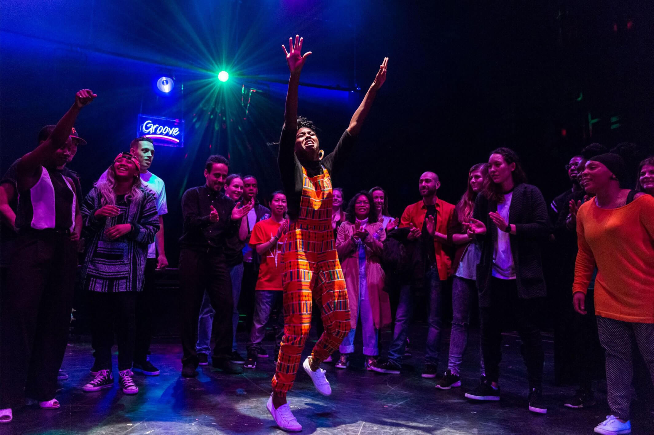 An African British female wearing orange dungarees with colourful rectangular patterns windmills her arms powerfully on the dance floor, the crowd that surround her clap their hands.