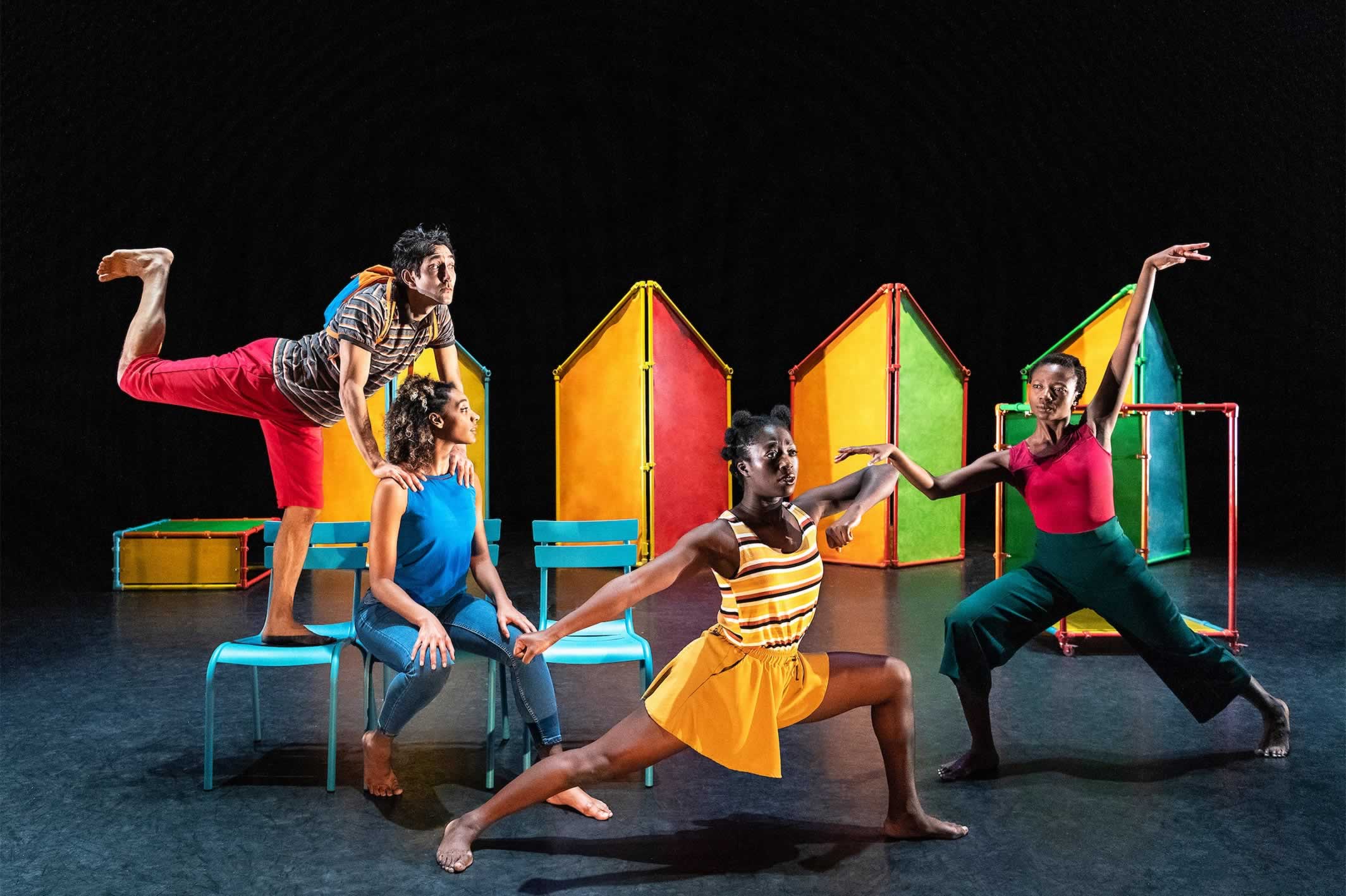 Four performers group together on set which combines different pastel colours: yellow, red, orange, light green, light blue, dark green. It looks a little bit like a kids’ playground.